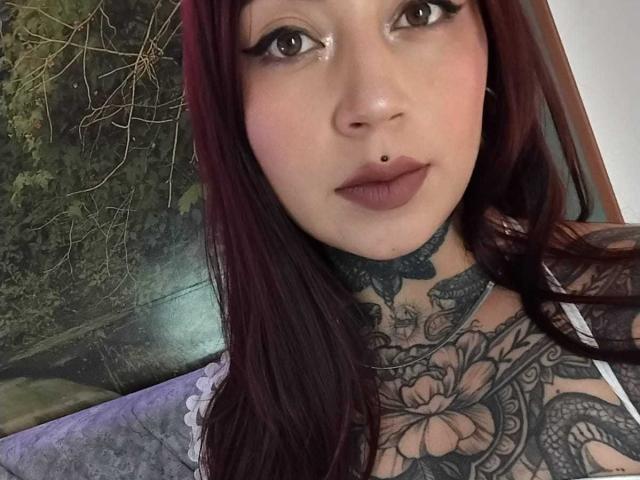 AmberKeller has dark red hair, black eyeliner, and a nose piercing. She's sporting tattoos on her neck and chest, and her tits are visible. Her lips are full.