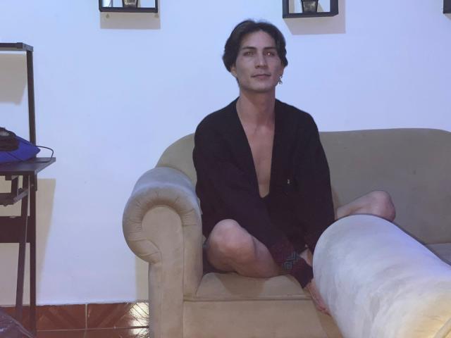 StevenHumps, shirtless, in a black jacket, sitting cross-legged on a beige couch, looking slightly to the right, with a relaxed, confident expression, and short, dark hair.