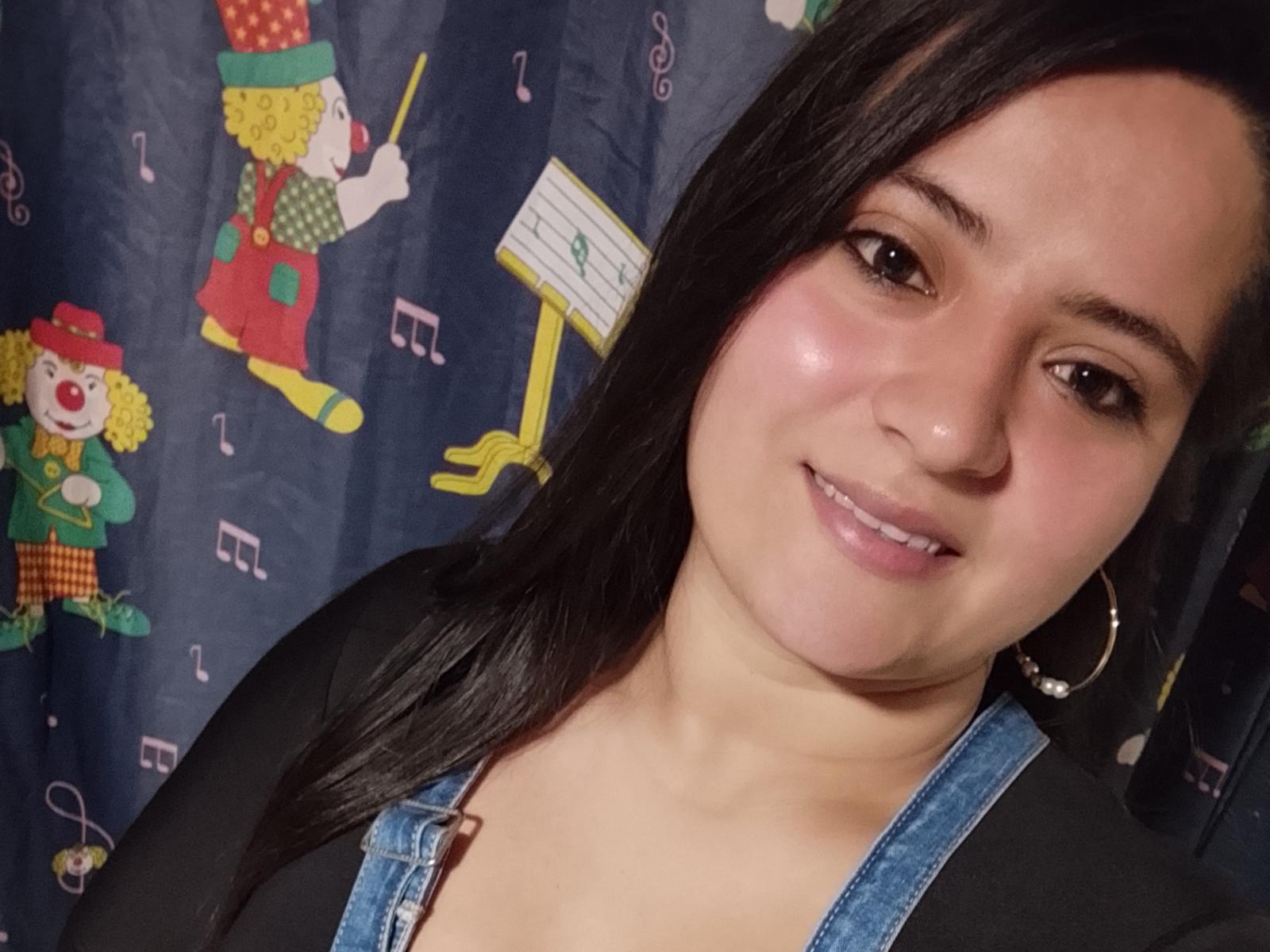 Nicoll69 has medium brown skin, dark straight hair, and is smiling with pink lips. She's wearing a black top and denim overalls, with hoop earrings, showing her tits.