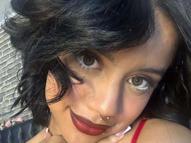 WendyJames has dark curly hair, green eyes, red lipstick, nose ring, and a slight smile. She's wearing a red top with white nails. Her ass is not visible.