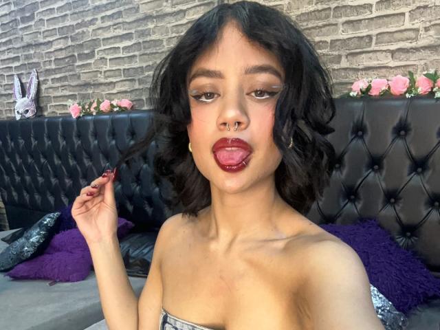 WendyJames has curly black hair, a nose ring, and dark red lipstick, sticking her tongue out while holding a lock of hair, wearing a strapless top, on a black tufted sofa.