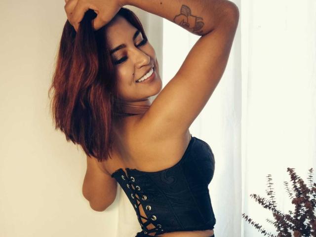 LucyDaniels with red hair, smiling, arms raised, wearing black lace-up corset, showing her tits, small tattoo on right arm, standing in bright room, fern in bottom right.