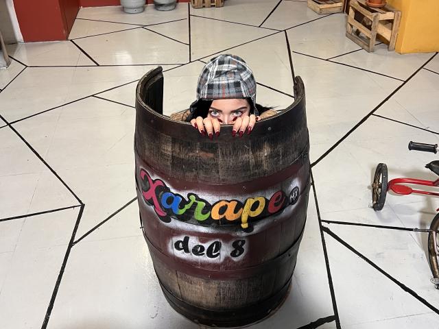 LillyTaylor is in a wooden barrel, peering out with red-painted nails and a plaid hat, her dark eyes looking forward. Her face is above the barrel's rim.