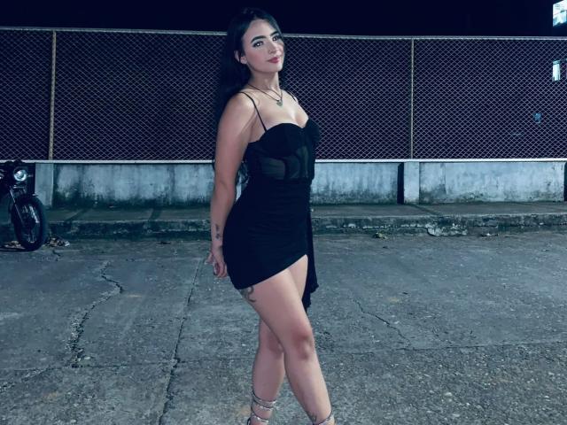KonanStar stands with her ass in a tight black dress, showing her tits, long black hair, tattoo on her right arm, and wearing strappy heels, on a cracked concrete floor.