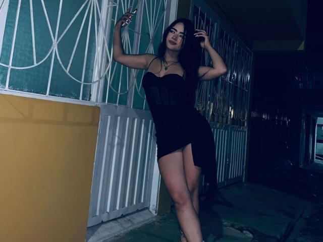 KonanStar, com longos cabelos pretos, vestindo um vestido preto, sem alças e transparente, com uma fenda alta, em frente a uma moldura de janela de metal branca, com os braços levantados, iluminação escura e melancólica..