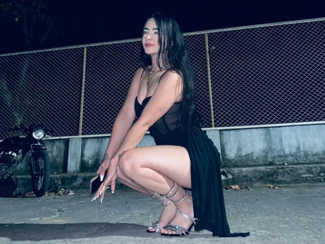KonanStar squats in a black dress with a high slit, showing her thighs, and wearing silver heels, against a chain-link fence at night, with a motorcycle on the left.