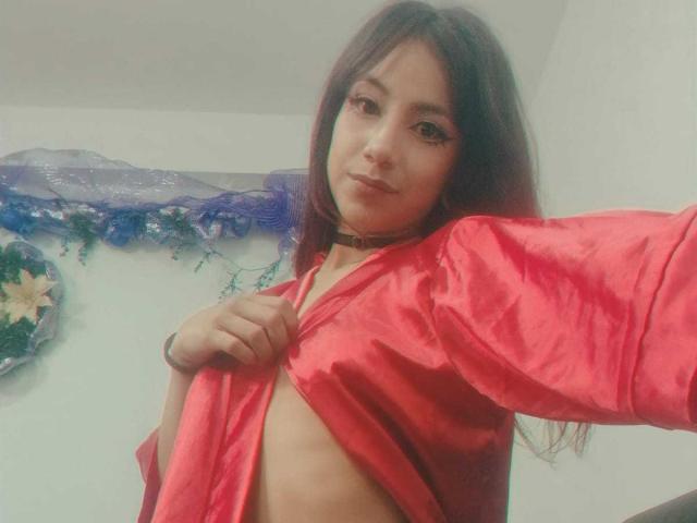 KarlaMiilan, com cabelos longos e escuros, vestindo uma camisa vermelha, levantando-a para exibir seus pequenos seios e sua barriga fina e plana. Ela usa um colar preto e tem um leve sorriso..