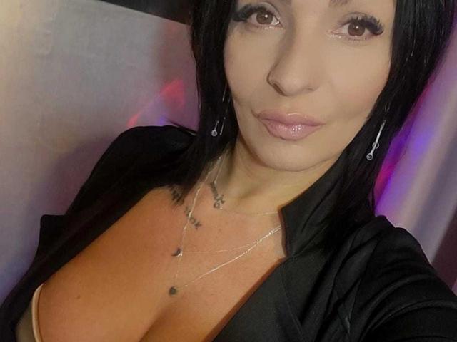 IsraKarma with black hair, pink lips, and large tits, wearing a black shirt and necklace, looking at the camera with a subtle smile.