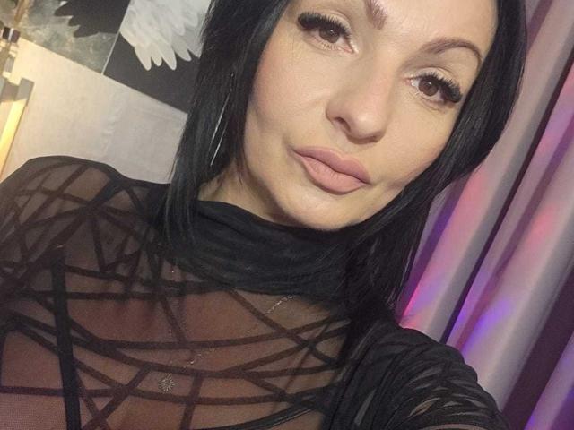IsraKarma with black hair, brown eyes, and pink lips, wearing a sheer black top with geometric patterns, showing her tits through the fabric. Her makeup is subtle and sexy.