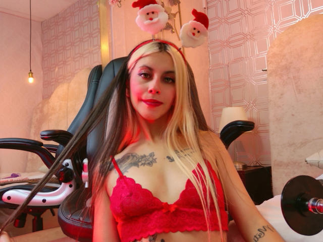 Yliria with long blonde and black hair, red lace bra, Santa headband, tattoos, and red lipstick, sitting in a black chair, smiling at the camera, red light on her face.