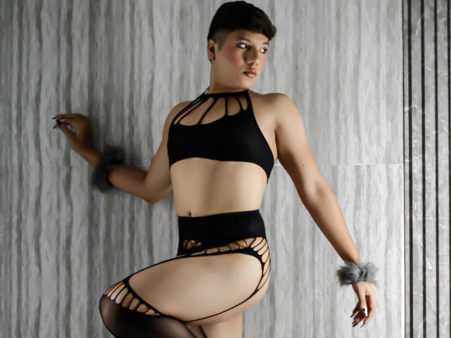 EllisBrown mit kurzen, braunen Haaren, trägt schwarze, trägerlose Unterwäsche, eine Strumpfband und graue Fellmanschetten. Sie steht an einer strukturierten Wand, blickt über die Schulter und zeigt einen sexy, kurvigen Hintern..
