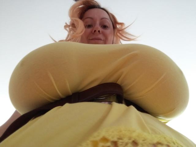 KarolyneOsyera with huge, yellow-dress-covered boobs, brown belt, and wavy, blonde hair, looking down at the camera with a slight smile. Low-angle, upward perspective.