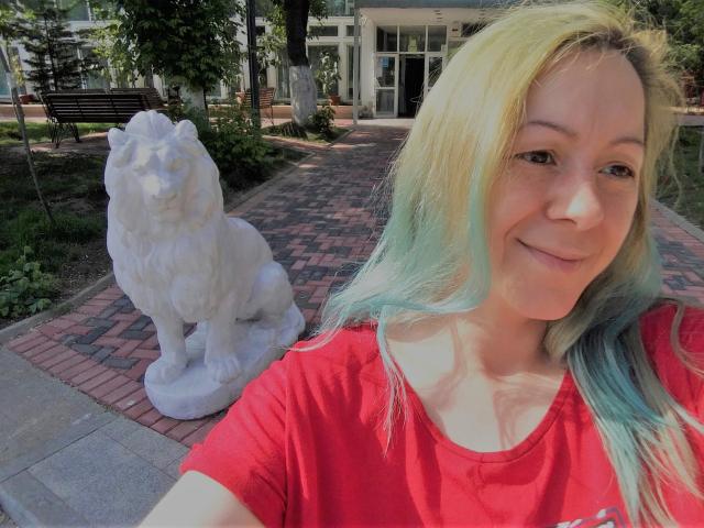KarolyneOsyera with long, blonde-green hair, smiling, wearing a red shirt, standing next to a white lion statue in a park. KarolyneOsyera's tits are small, and she's cute.