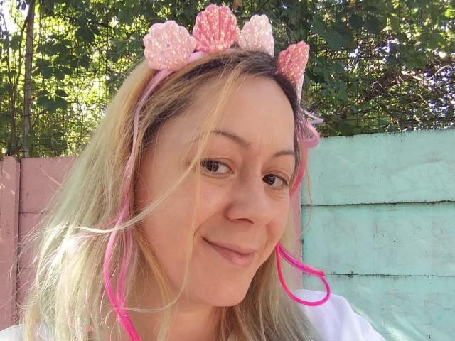 KarolyneOsyera has blonde hair, pink headband with pink polka dots, pink ear loops, white shirt, light skin, small nose, subtle smile, and green leafy background in sunlight.