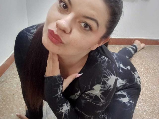 Gaiax69 with pouty red lips, long dark hair in a ponytail, wearing a black, white splatter-patterned long-sleeve top, kneeling on a speckled floor, touching her neck.