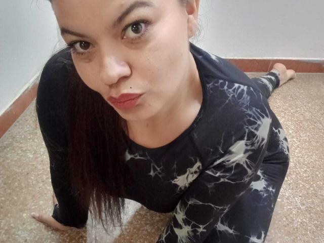 Gaiax69 is on all fours, wearing a black, white, and gray patterned top and matching pants, with a small mole above her red-lipped, upturned nose, and long brown hair.