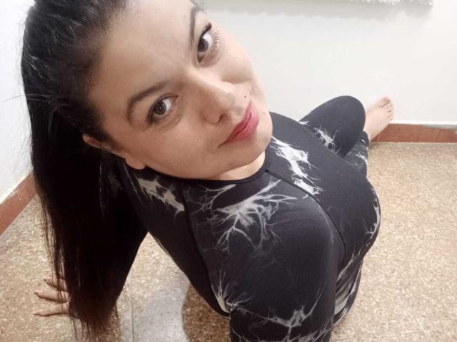 Gaiax69 with long black hair, looking up, wearing a black tie-dye top, sitting on a brown-speckled floor, smiling, slightly tilted head, showing her nose and brown eyes.