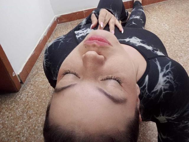 Gaiax69 is lying down with eyes closed, wearing a black and white patterned top, hands on chest, brown carpet, and brown wood trim in the background.