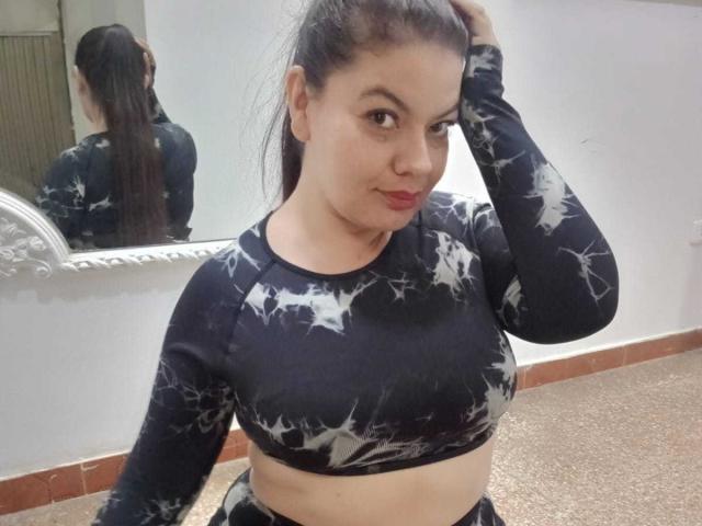 Gaiax69 with dark hair in a ponytail, wearing a black star-patterned crop top, showing her tits, hand in hair, mirror reflection, and red lipstick, standing in a room.