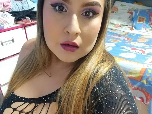 SweetAfhrodita with long blonde hair, purple eyeshadow, and pink lipstick, wearing a black, starry, cut-out top, showing her tits, in a bedroom with colorful bedspread.