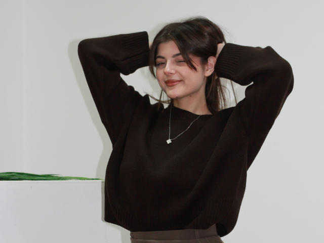 StellaLewin with arms raised, wearing a dark brown sweater, smiling, small tits, brown hair in a ponytail, necklace with a heart-shaped pendant, standing in front of a white wall.