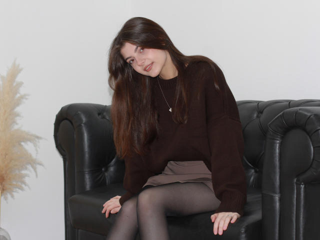 StellaLewin with long brown hair, brown sweater, and black tights, sitting on a black leather couch, showing her ass, looking down with a slight smile.