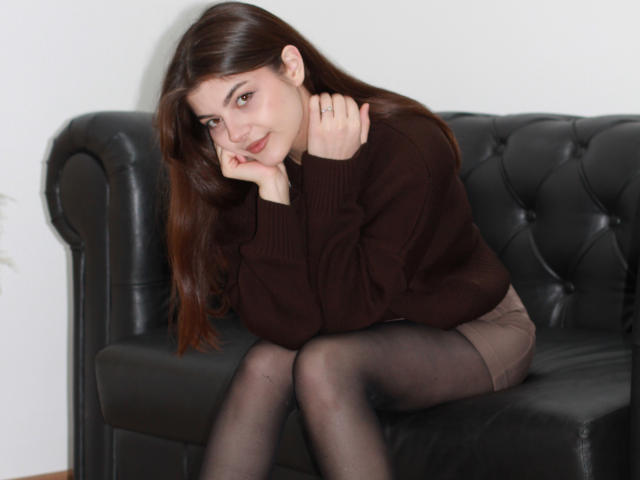 StellaLewin with brown hair, sitting on a black leather couch, wearing a brown sweater, black pantyhose, and a brown skirt, resting her chin on her hand, smiling.