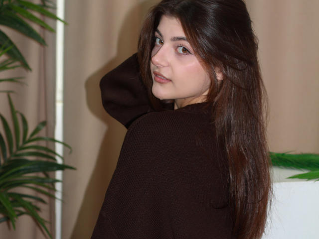StellaLewin with long brown hair, brown eyes, and pink lips, wearing a dark brown sweater, looking over her shoulder with a slight smile, hand in hair. Potted plant left.