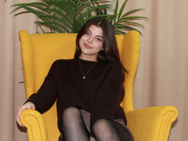 StellaLewin with long brown hair, black sweater, and black tights, sitting in a yellow chair, smiling slightly, wearing a silver necklace, hands on arms, green plant behind.