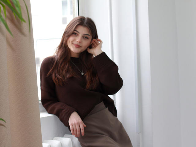 StellaLewin with brown hair, brown sweater, and brown skirt, sitting by window, smiling, hand on cheek, necklace, soft light, white walls, green plant, casual, cute, relaxed.