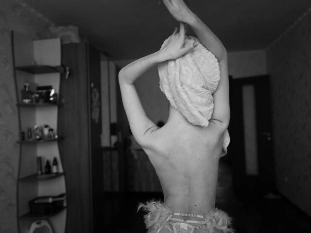 BlondiAngel-hot with a towel wrapped around her head, arms raised, exposing her bare back and a frilly bottom, standing in a dark, empty room. Black and white.