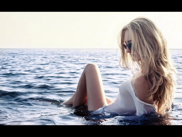 Consento-hot with blonde hair, wearing sunglasses and a wet white shirt, sits in dark blue ocean water, legs slightly apart, looking downward. Her tits are visible through the shirt.