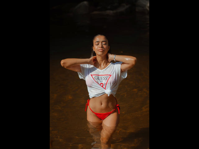Consento-hot with wet white tee, tied at front, showing small tits, red string bikini bottom, hands behind head, standing in shallow brown water, sunlit, eyes closed, serene.