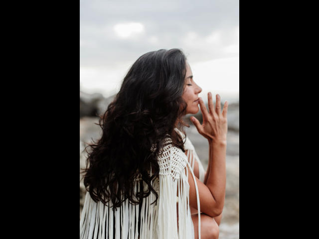 Consento-hot with wavy black hair, wearing a white fringe shawl, eyes closed, hands near face, standing against a blurry background. Soft, natural light.