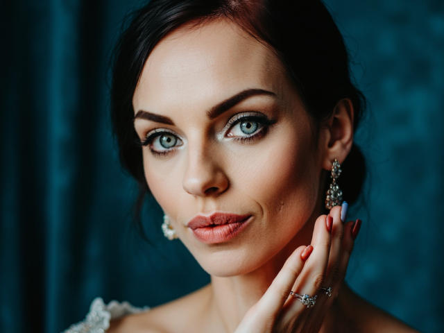 CassieBest has blue eyes, dark hair, and pink lipstick. She's wearing earrings and a ring, with red nails touching her chin. Her skin is fair, and she's looking at the camera.