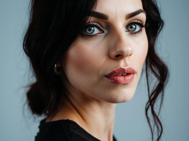 CassieBest has blue eyes, dark wavy hair, and pinkish-red lipstick. Her face is slightly turned to the right with a subtle, pouty expression. She's wearing a black top.