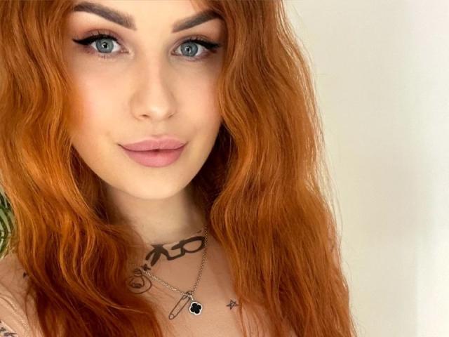 Close-up of KiraNeeww with long, wavy red hair, fair skin, and blue eyes. She has bold, black eyeliner and pinkish lips. She wears a necklace with a black cross and a small pendant. A tattoo with black script is visible on her chest. Her eyebrows are thick and well-defined. The light is even, highlighting her hair's texture and her smooth skin. The image is sharply focused on her face, with no visible background. Slight smile on her lips. Overall, the composition emphasizes KiraNeeww's facial features and hair.