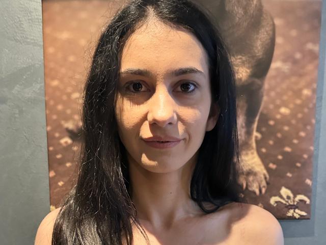 SilviaVanda has long black hair, brown eyes, and a slight smile. She's bare-shouldered with a tan skin tone and a dog's blurred ass in the background.