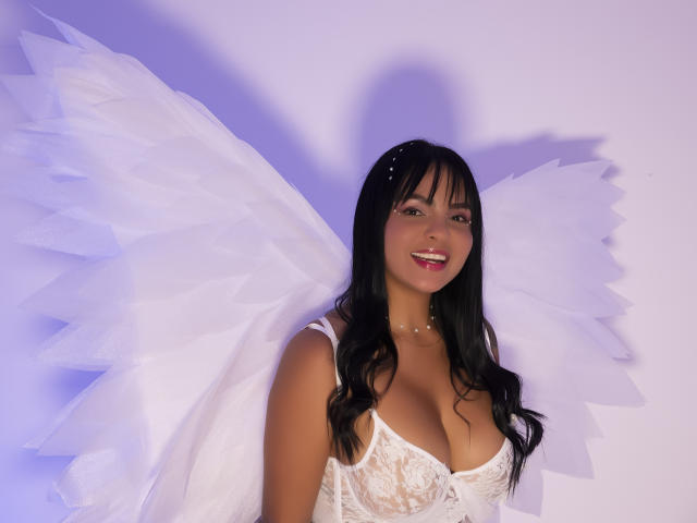 JessicaFiore met witte engel vleugels, zwart haar en een witte kantjurk, die haar grote borsten laat zien en die lacht. Ze draagt een parelketting en heeft een kleine haarspeld..