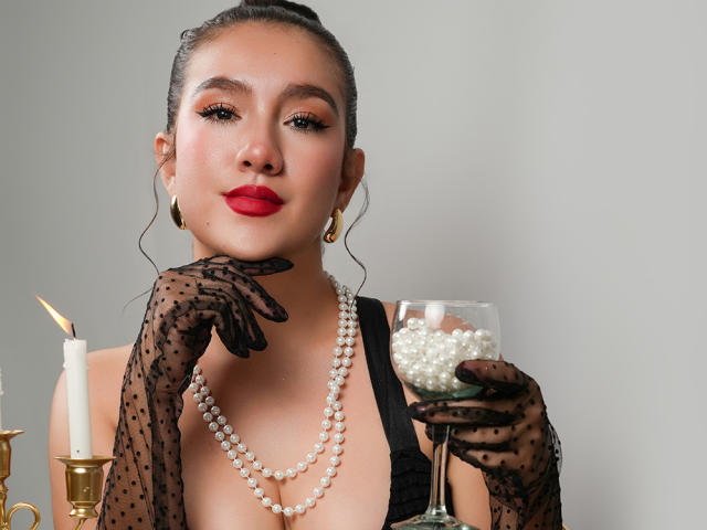Zhara mit rotem Lippenstift, schwarzen Handschuhen mit Punkten, einer Perlenkette, einem Glas mit Perlen, goldenen Ohrringen und einer weißen Kerze in einem Messing-Halter, blickt mit einem selbstbewussten Lächeln in die Kamera..