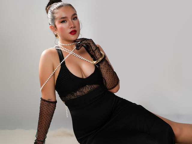 GagingZhara in a black lace dress, holding a pearl necklace, with polka-dot gloves, red lipstick, and her tits showing. She's sitting, looking sexy, with her ass slightly raised.