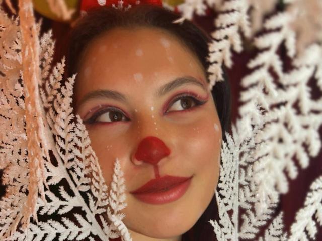 DominicTheOne has a red nose, pink eyelids, and is surrounded by white, fluffy, Christmas-like branches. She's smiling with a subtle glow on her face.