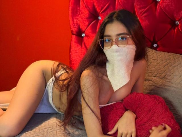 SweetlLove69 with long brown hair, glasses, and a white mask, lying on a bed in white panties, showing her tits and ass, holding a red pillow, against a red headboard.