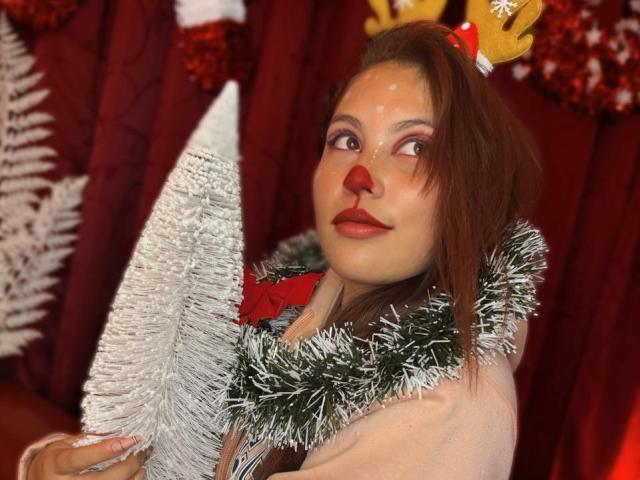 DominicTheOne has red nose makeup, brown hair, and antlers, holding a silver Christmas tree. Wearing a silver tinsel scarf and pink top, with red lipstick.