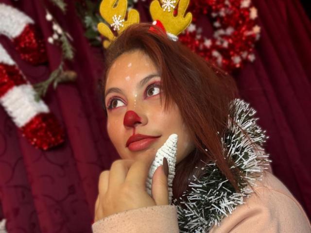 DominicTheOne has red nose, Christmas antlers, pink makeup, and brown hair, holding silver tinsel, wearing pink sweater, with red and white decor in background.