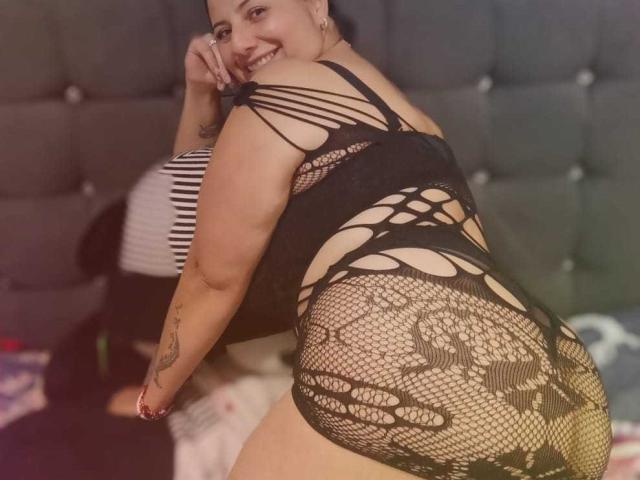 AngeleRoux, curvilínea com um traseiro grande, vestindo um vestido de renda preta com amarração, ajoelhada em uma cama, sorrindo para a câmera, mostrando tatuagens em seu braço..