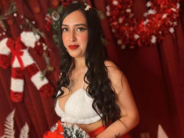 Chloey69 with long black hair, red lipstick, white fur bra, holding red and white ornament, red curtain, red and white decorations, tattoo on chest, looking at camera.
