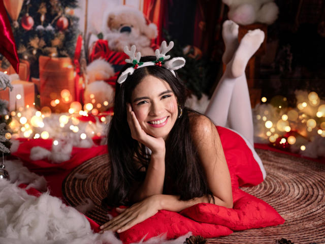 MadisonSmitt, lying on red pillow, wearing white thigh-highs, red dress, Christmas headband, smiling, hand on cheek, legs up, cozy Christmas setup, candles, Santa, festive decorations.