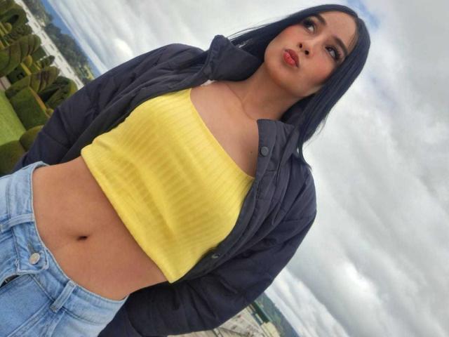 ValentinaRisso tiene el pelo azul, lleva un top corto amarillo con textura, que deja ver sus pechos, y una chaqueta azul, con jeans azules, y está de pie afuera bajo un cielo nublado..