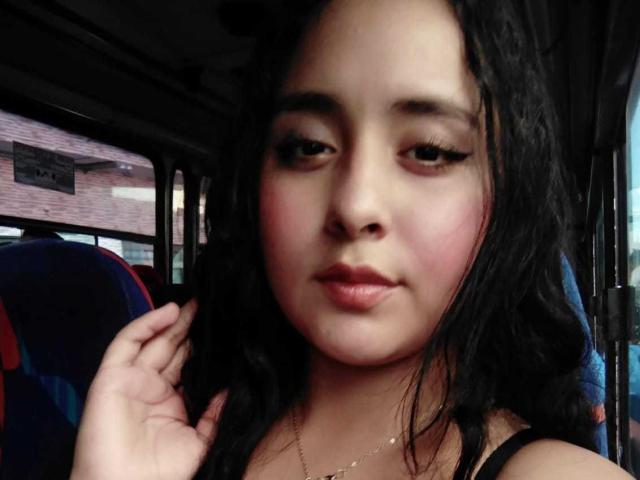 ScarlettZ has wavy black hair, pink blushed cheeks, and is looking at the camera with a slight smile, wearing a black top, in a bus. Her hand touches her hair.